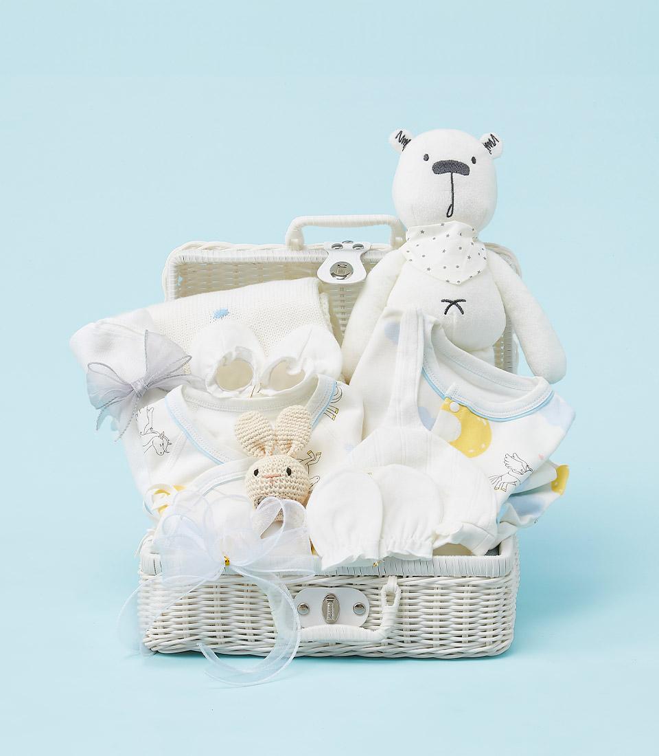 A white wicker basket filled with baby essentials, including a plush bear, clothes, a rattle, and a blanket.