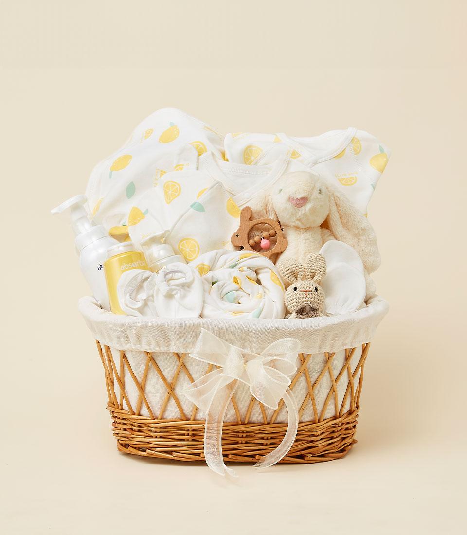 A wicker gift basket filled with baby items, including white and yellow Lemon Sunshine-patterned clothing, a stuffed bunny toy, rolled-up blankets, baby lotion bottles, and a small wooden rattle. The basket is decorated with a sheer white ribbon bow.