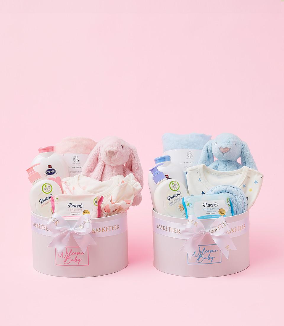 Two round gift baskets sit on a pink background, each serving as a Gentle Baby Welcome Gift Box. They contain baby items like lotion, soap, clothing, a plush bunny, and a bib. The left basket has a pink theme with a pink bunny, while the right basket features blues with an adorable blue bunny.
