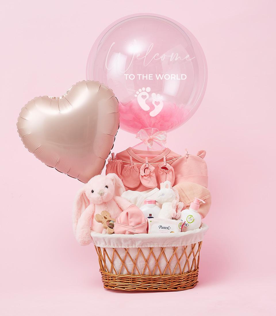 A charming baby girl welcome basket filled with pink items, including a stuffed bunny, baby clothes, and baby care products. Two balloons are attached: one heart-shaped and the other a clear bubble with pink feet and 