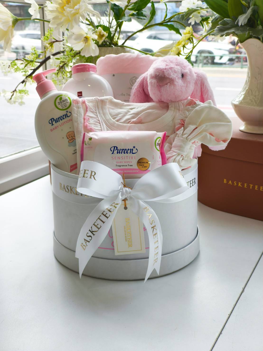 Newborn baby gift basket with Pureen baby lotion, baby wipes, baby clothes, blanket, and plush pink bunny in a white round box with satin ribbon.
