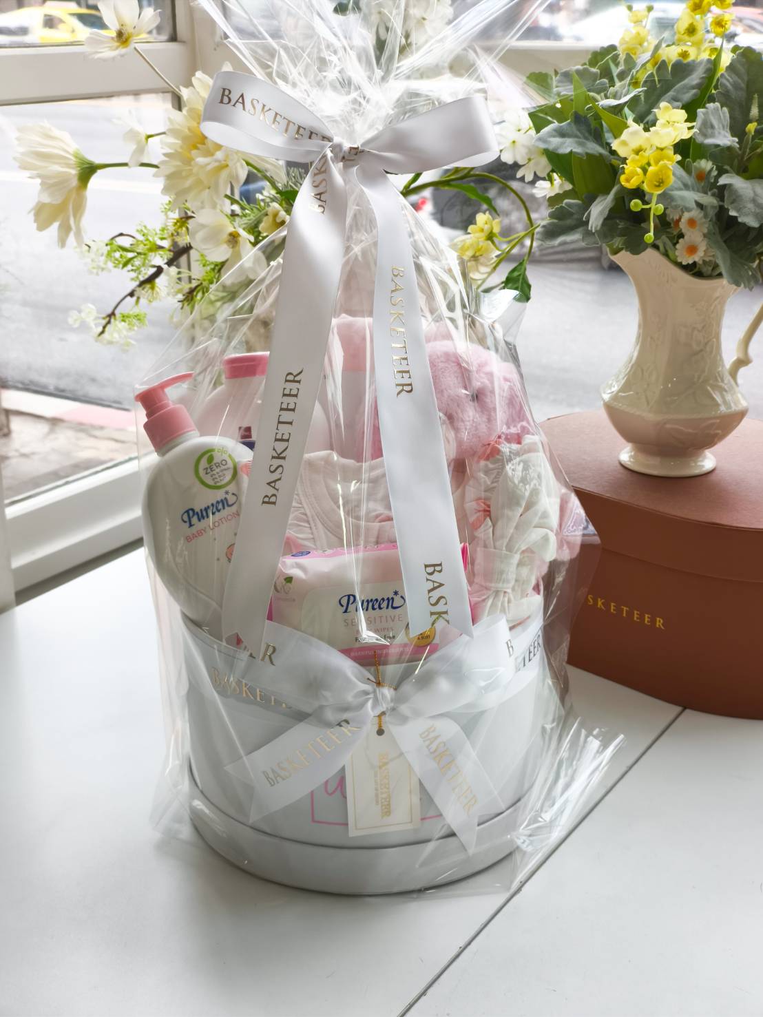 Newborn baby gift basket with Pureen baby lotion, baby wipes, baby clothes, blanket, and plush pink bunny in a white round box with satin ribbon