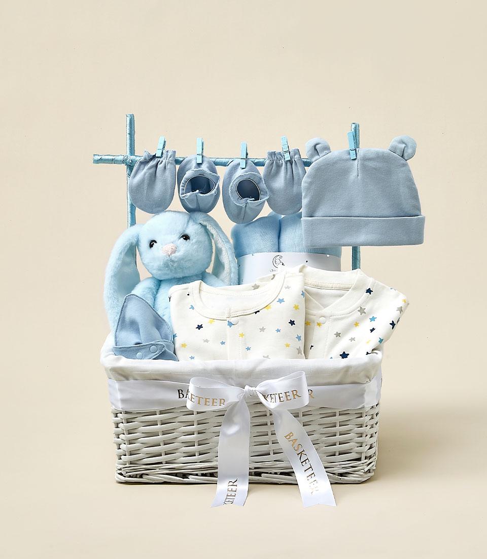 A gift basket with Baby's First Essentials includes a stuffed blue bunny, baby hats, mittens, booties, and onesies decorated with stars. The items are predominantly blue and white, and a ribbon around the basket is labeled 