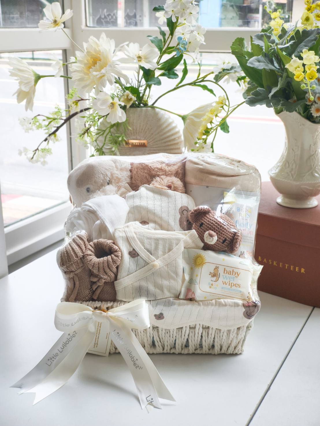 Luxury newborn baby gift basket featuring baby clothes, knit socks, wipes, plush toy bear, and essentials in a white woven basket with satin ribbon.