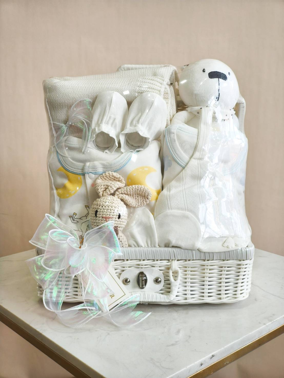 Luxury newborn baby gift basket with white baby clothes, knit blanket, plush bear, crochet bunny, and essentials in a white wicker basket with satin bow.