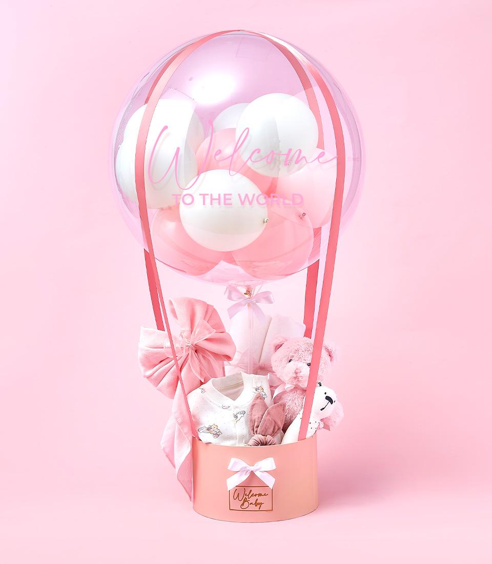 A pink-themed baby gift arrangement featuring a transparent balloon with white and pink balloons inside, displaying the text 