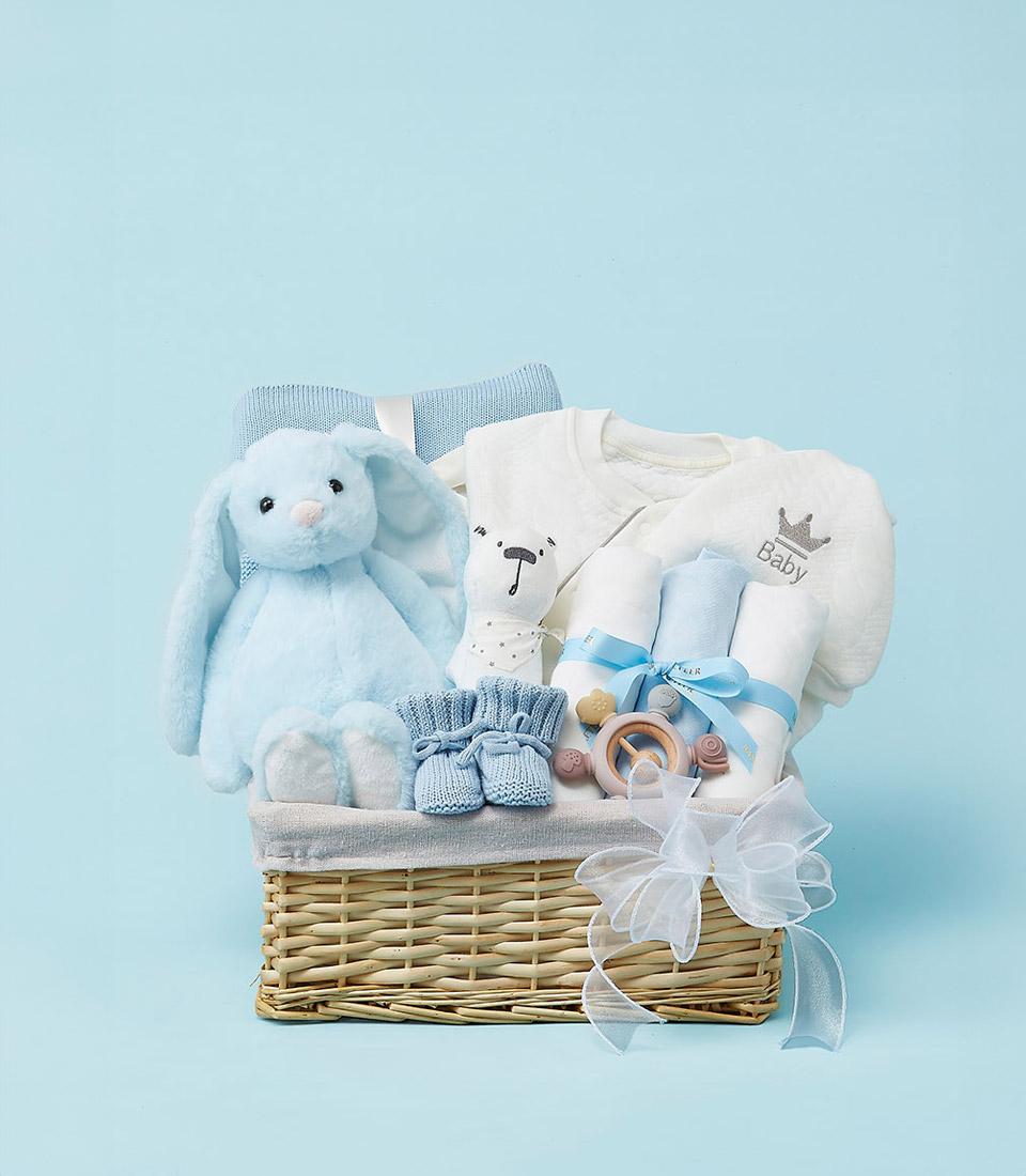 A cute newborn baby welcome bundle gift arrives in a wicker basket overflowing with baby gifts: a blue stuffed bunny, knitted booties, a folded blue knit blanket, a white onesie with 