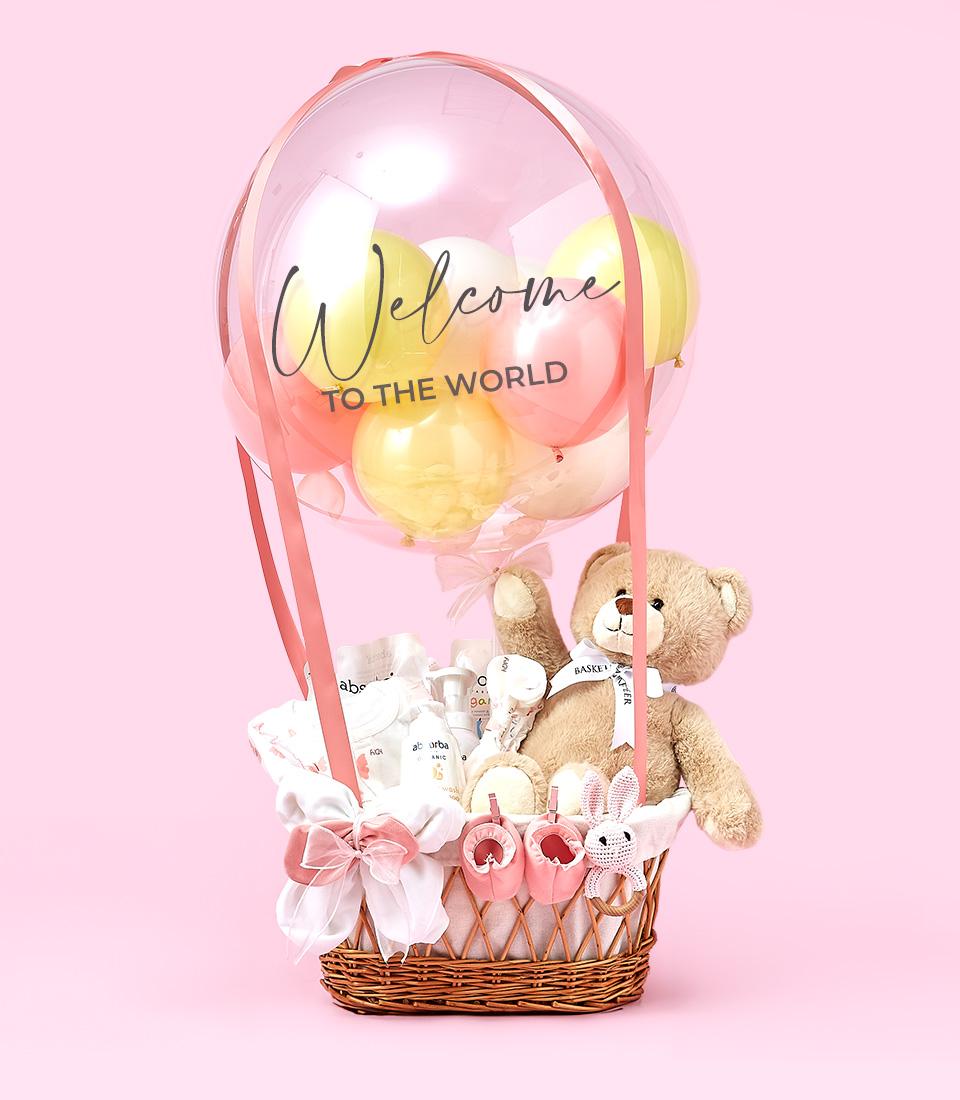 A baby gift basket contains plush toys, baby clothing, and baby care products. It is adorned with a large transparent balloon with smaller pink, yellow, and white balloons inside. The balloon is labeled 