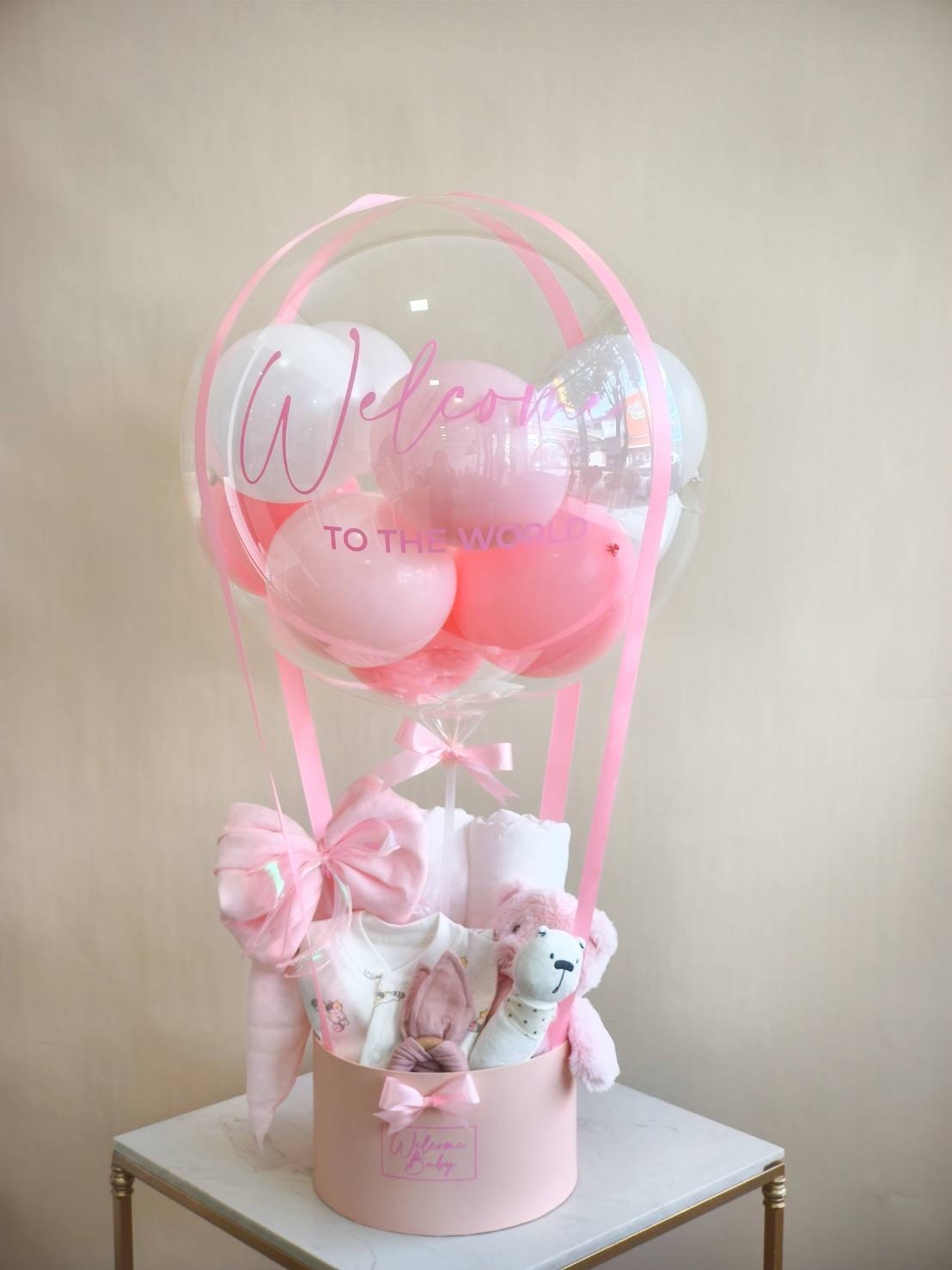 Baby gift balloon box with pink and white balloons, teddy bear, baby clothes, and essentials, decorated with satin ribbons and “Welcome to the World” message.