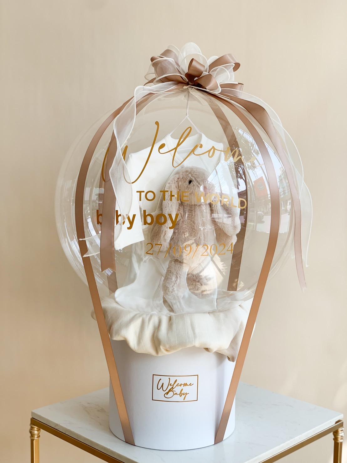 A decorative hot air balloon-shaped gift features a clear balloon with gold lettering that reads "Welcome to the world baby boy 27-07-2024." Inside the balloon, there's a plush bunny. It sits in a white basket with a white ribbon and bow, placed on a small table.