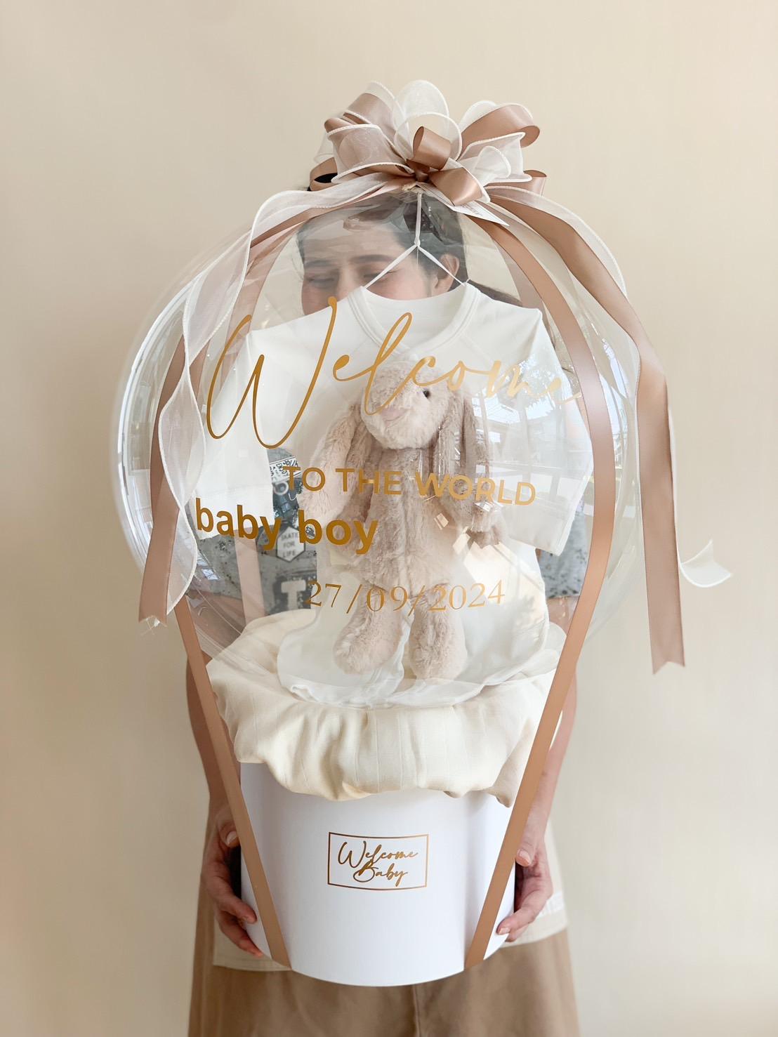 A person holds a clear balloon with "Welcome to the World, baby boy" and "27/09/2024" written on it. Inside the balloon is a stuffed teddy bear, reminiscent of a Happy Birthday balloon. The balloon is adorned with beige ribbons and attached to a white box with a "Welcome Baby" label.