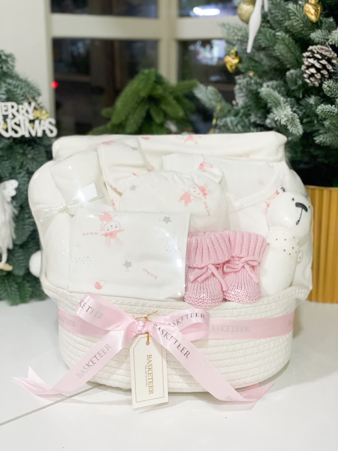 A charming newborn gift basket by Basketeer, featuring baby essentials such as soft clothing, a plush toy, pink knitted booties, and cozy blankets, elegantly presented in a white basket adorned with a pink ribbon, set in a festive Christmas-themed backdrop.