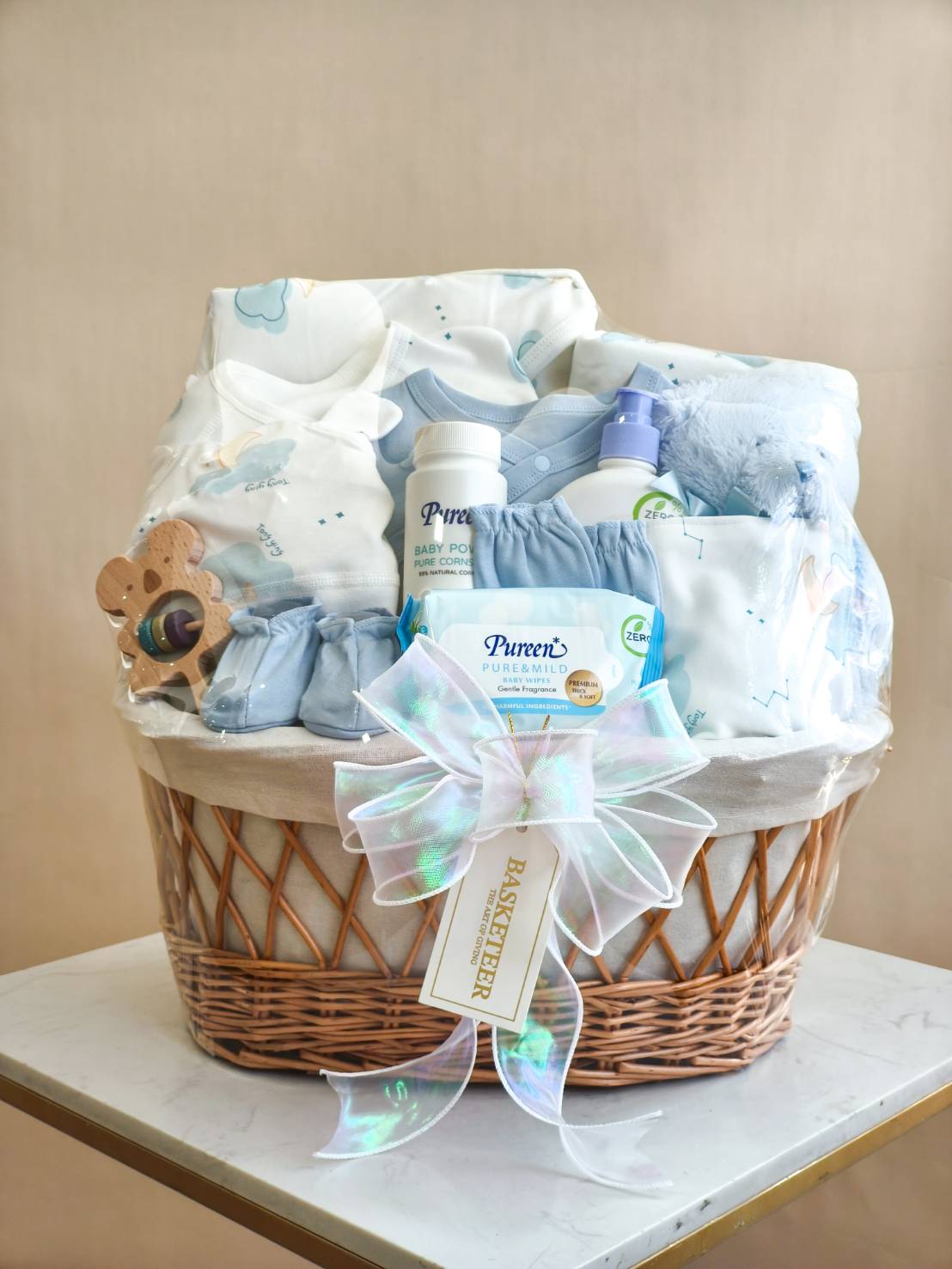 Luxury baby gift basket with blue newborn clothes, Pureen baby care, wipes, plush toy, and wooden rattle, decorated with premium ribbon