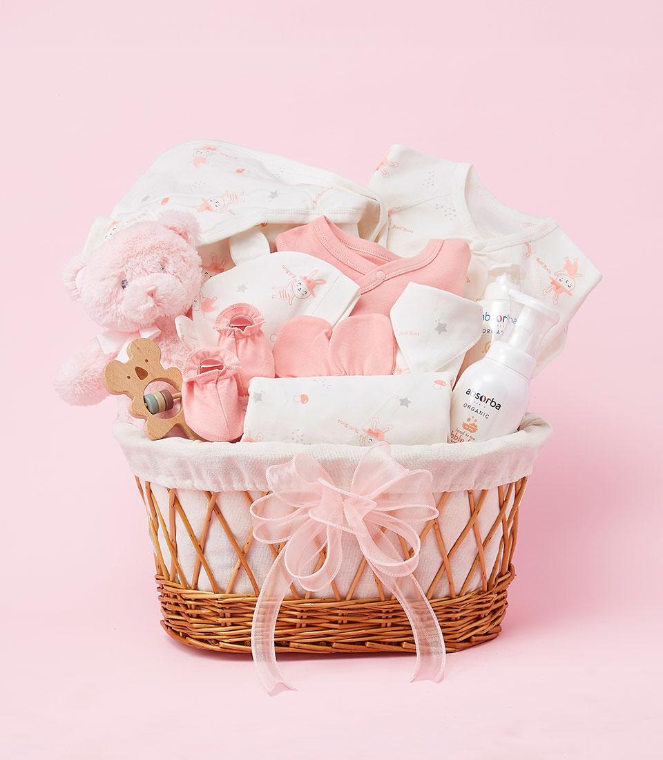 A pink wicker basket filled with baby essentials, including a plush bear, clothes, a rattle, and baby care products.