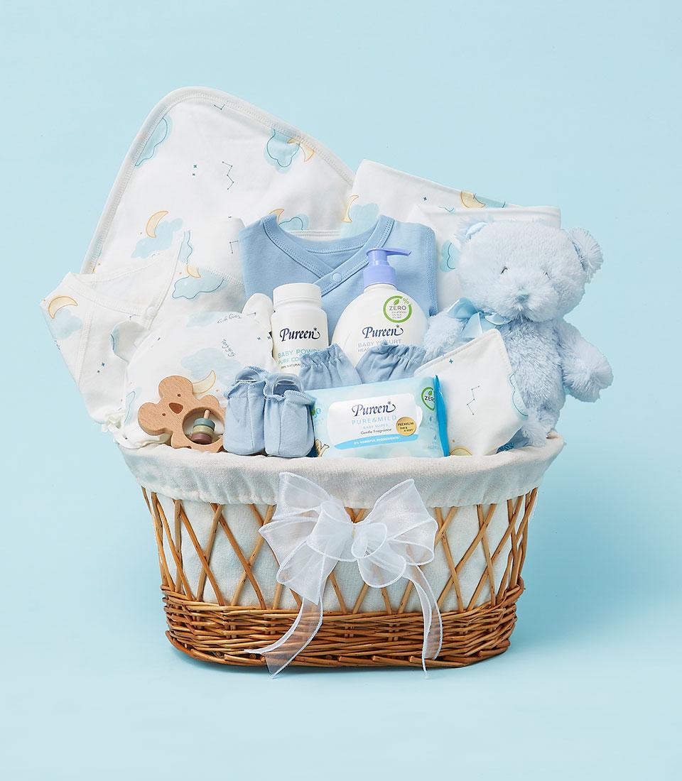 A blue wicker basket filled with baby essentials, including a plush bear, clothes, a blanket, and baby care products.