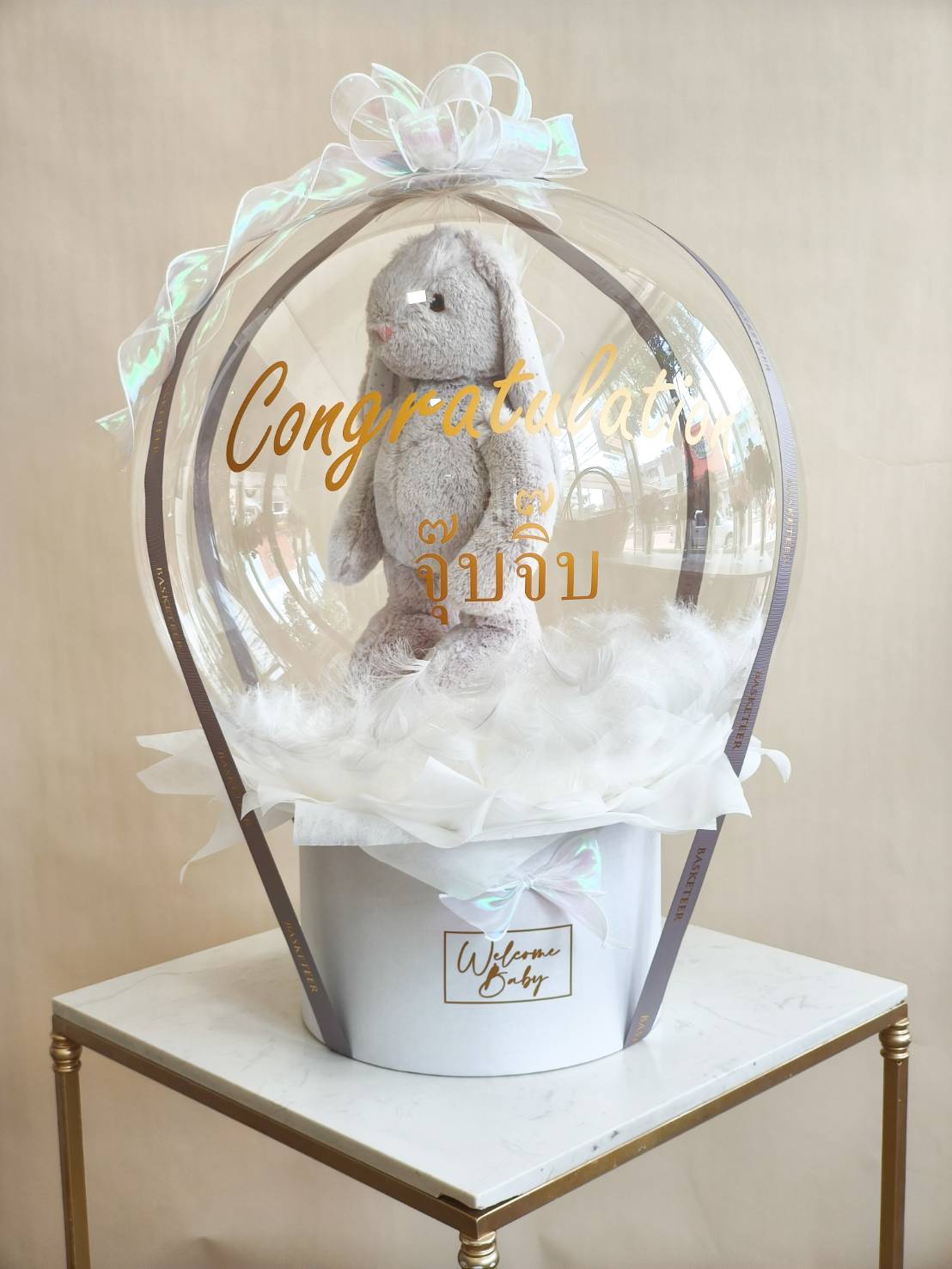 Luxury bubble balloon gift box with bunny plush toy inside, decorated with white fluffy base, ribbon, and Congratulations message.