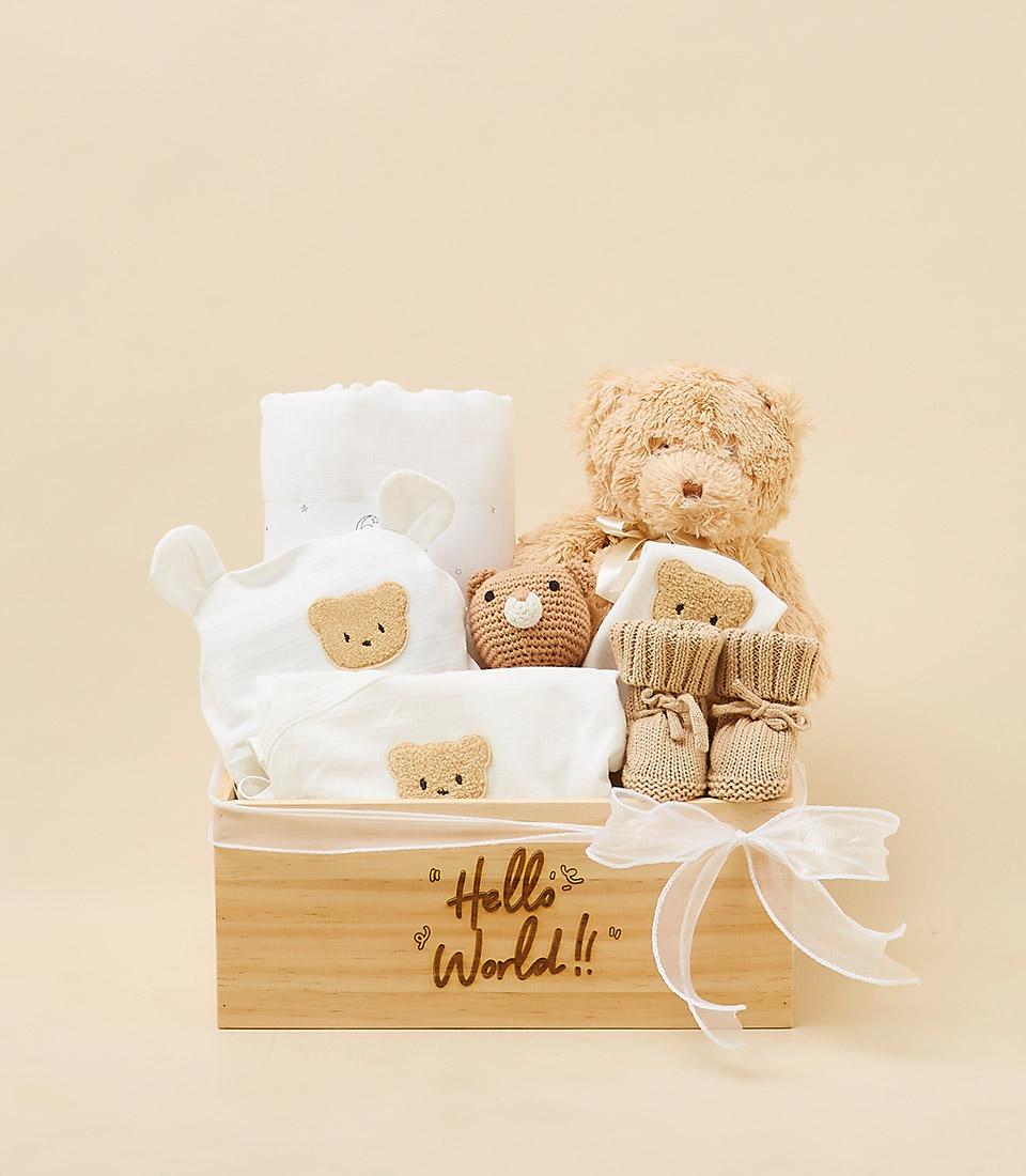 A wooden gift basket filled with baby essentials, including a teddy bear, clothes, a blanket, and a rattle