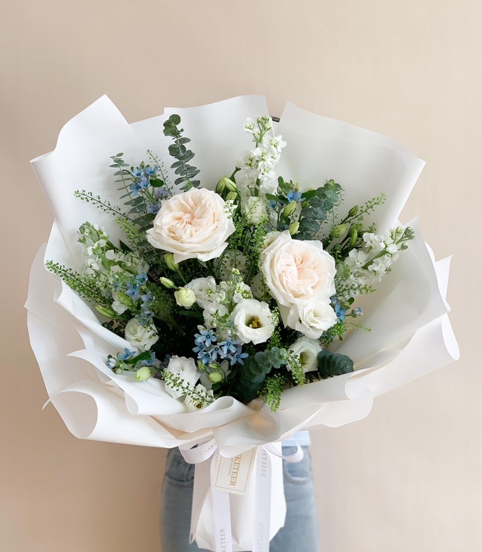 A large, elegant bouquet of flowers featuring pale pink roses, white lisianthus, blue delphiniums, and various green foliage, wrapped in white paper. The bouquet is being held against a plain, beige background.