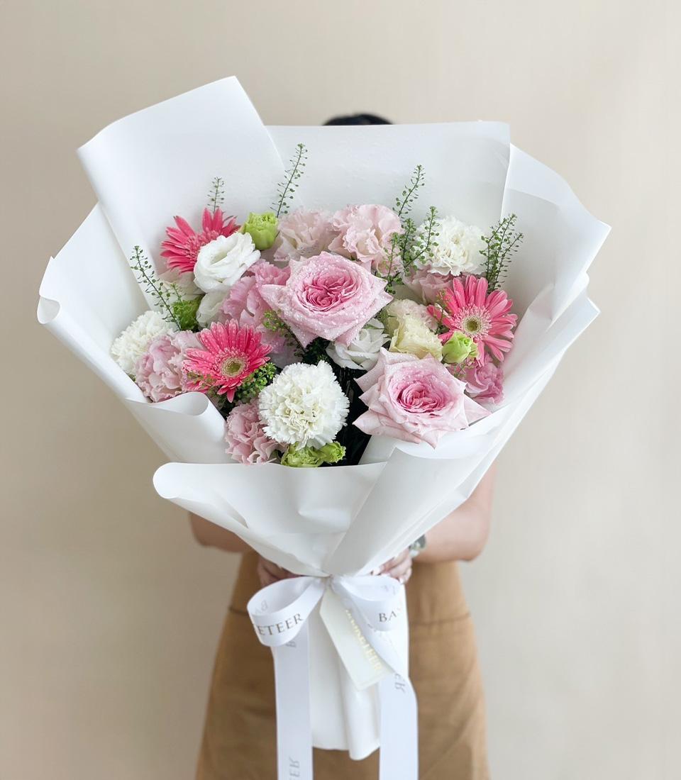 A person in a light brown apron holds a large bouquet of flowers wrapped in white paper. The bouquet features pink roses, pink gerbera daisies, white carnations, and greenery. A white ribbon with text is tied around the base of the bouquet.
