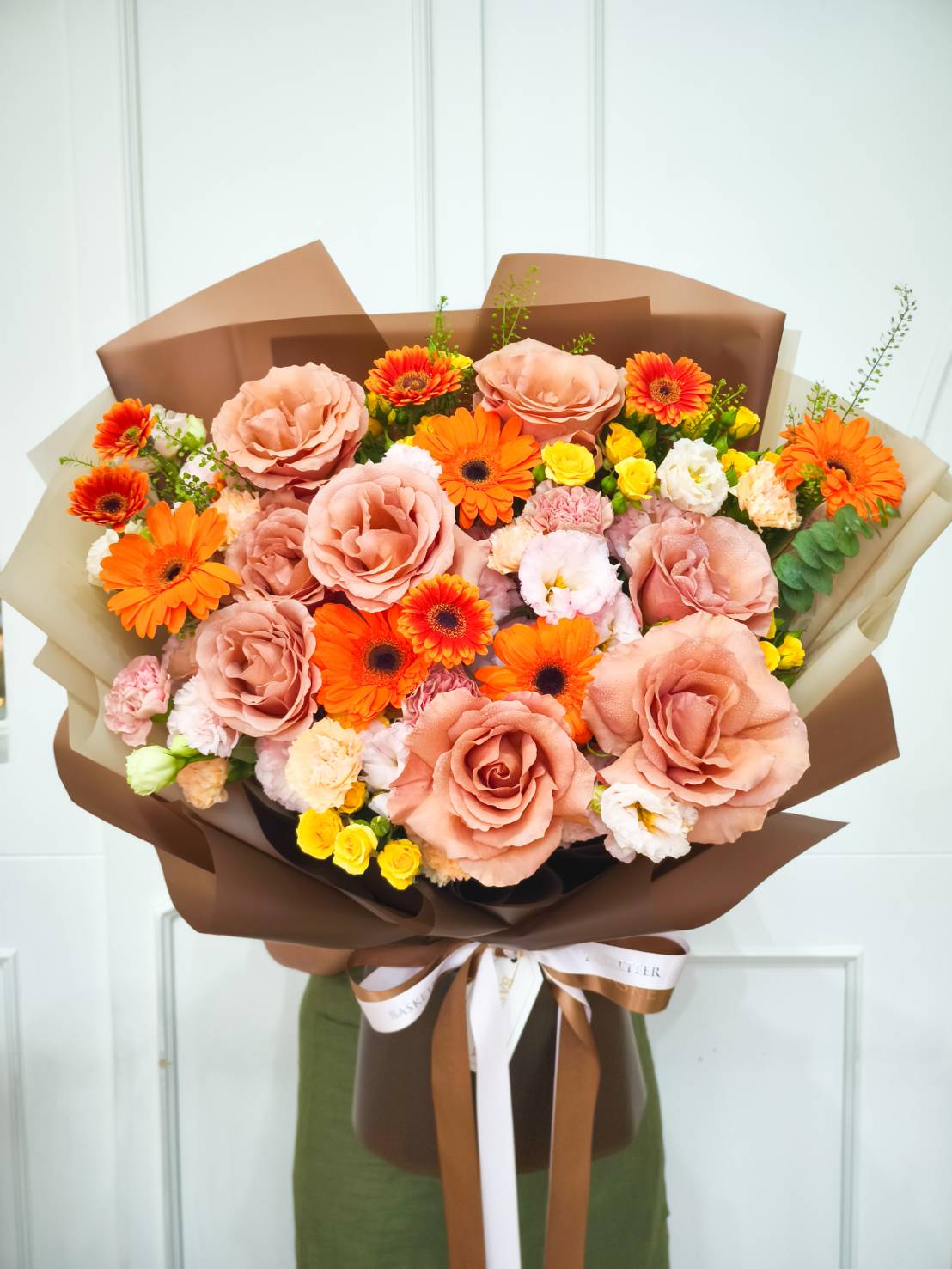 Luxurious bouquet of mocha roses, orange gerberas, and mixed blooms wrapped in brown paper with satin ribbons.