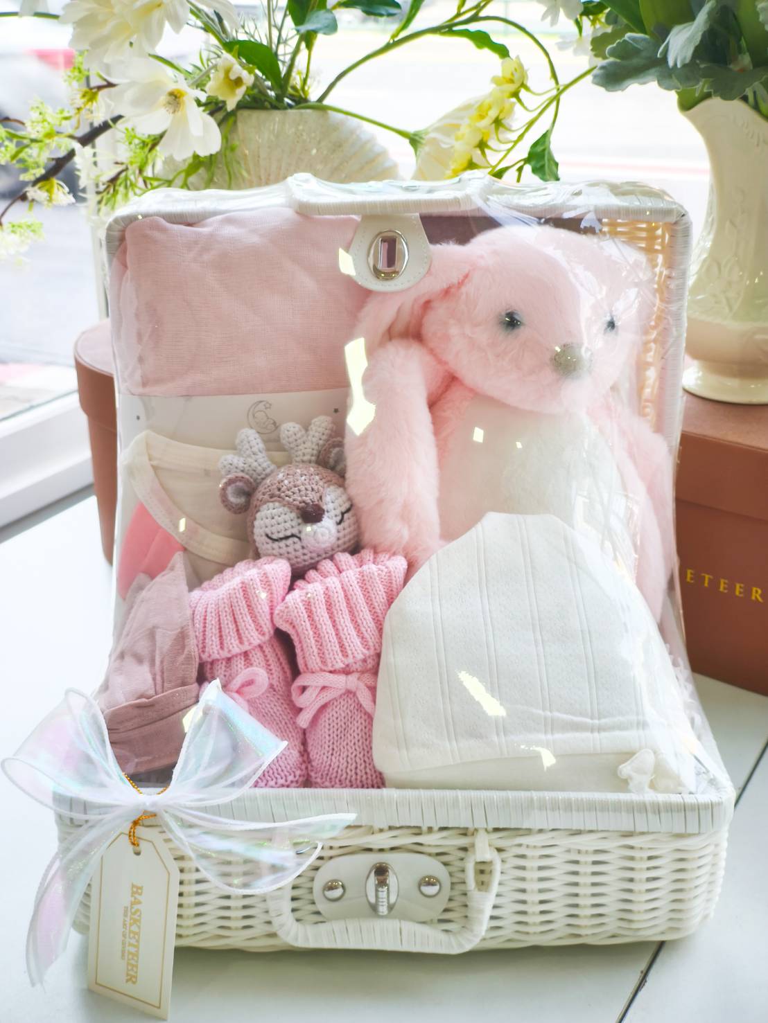 Premium baby gift hamper in white wicker basket with pink baby clothes, knitted socks, hat, blanket, plush bunny, and crochet toy.
