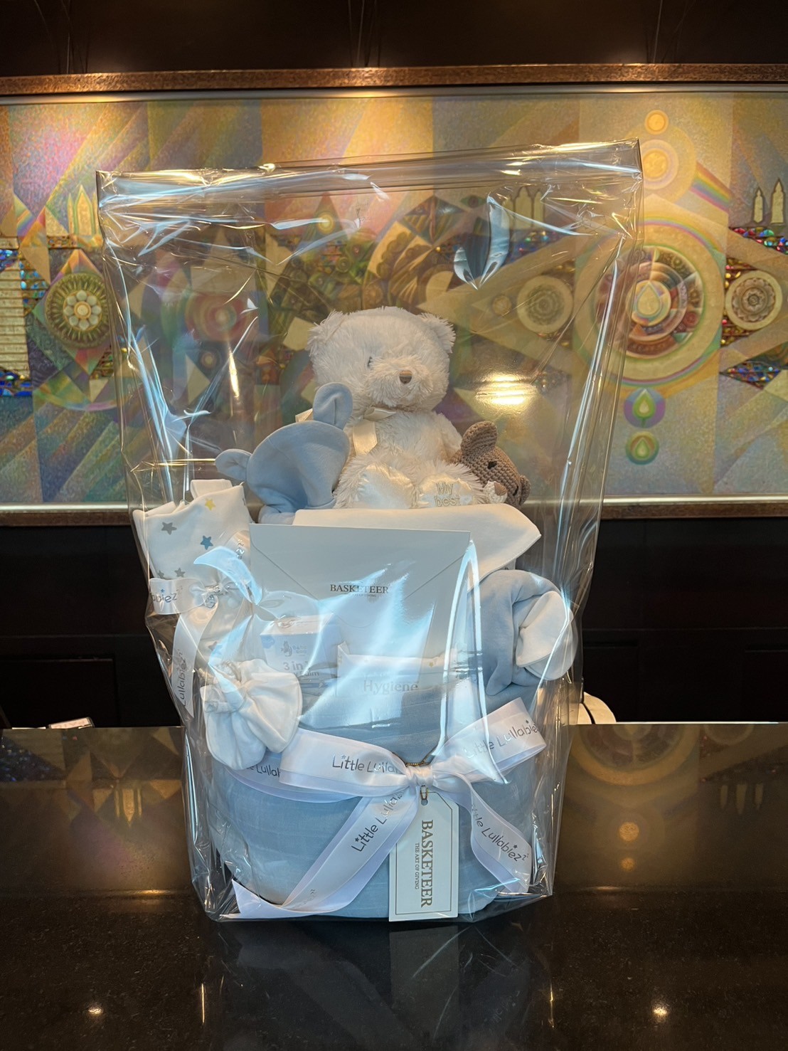 A gift hamper wrapped in clear plastic features Blue Skies and Teddy Bears, with baby clothes in blue and white and a grey soft toy, all set on a dark worktop against a colourful mosaic wall.