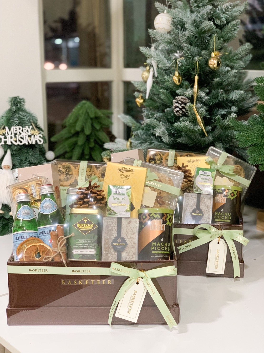 Gourmet gift hamper featuring coffee, herbal tea, chocolate, and sparkling water in a brown box with festive ribbon and accents.