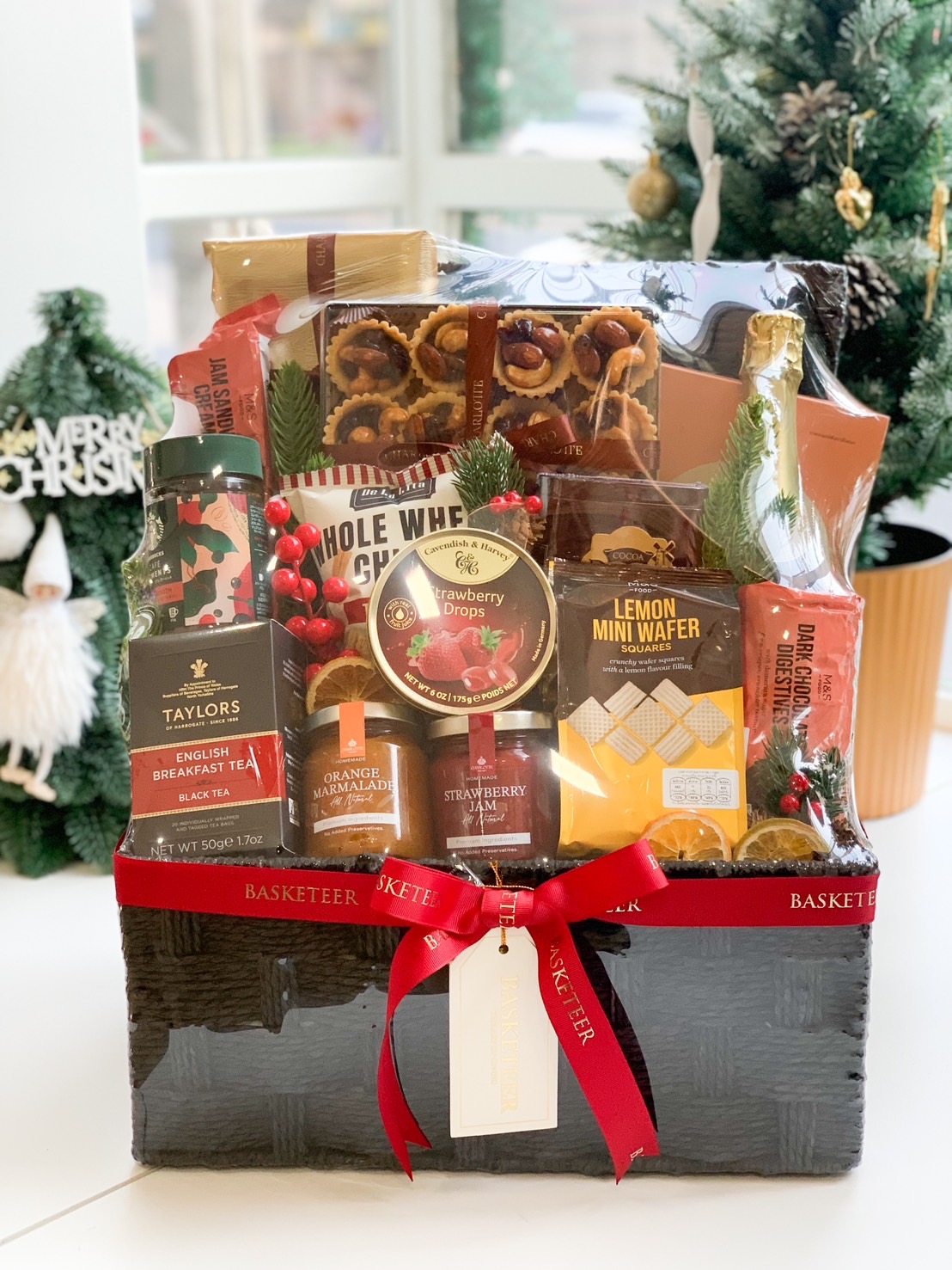 A black wicker gift basket filled with tea, cookies, wafers, strawberry jam, orange marmalade, and festive red decorations, tied with a red ribbon.