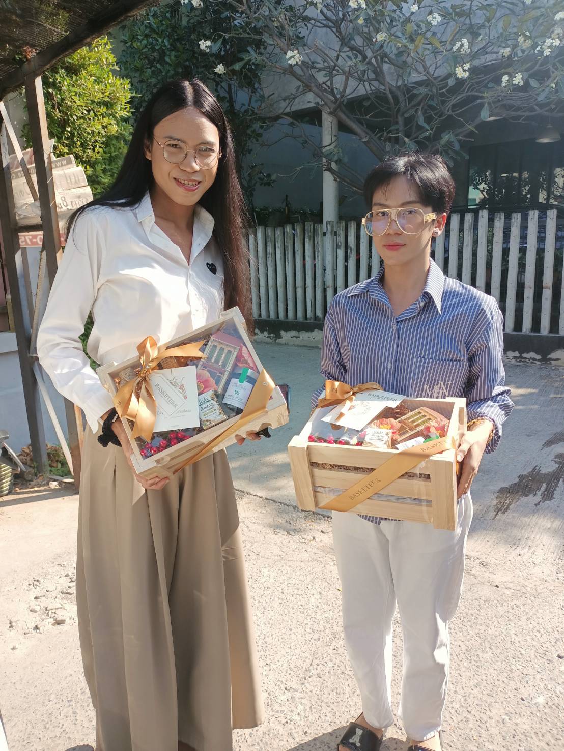 Two individuals holding elegant wooden gift boxes filled with premium snacks and treats, adorned with golden ribbons by Basketeer.