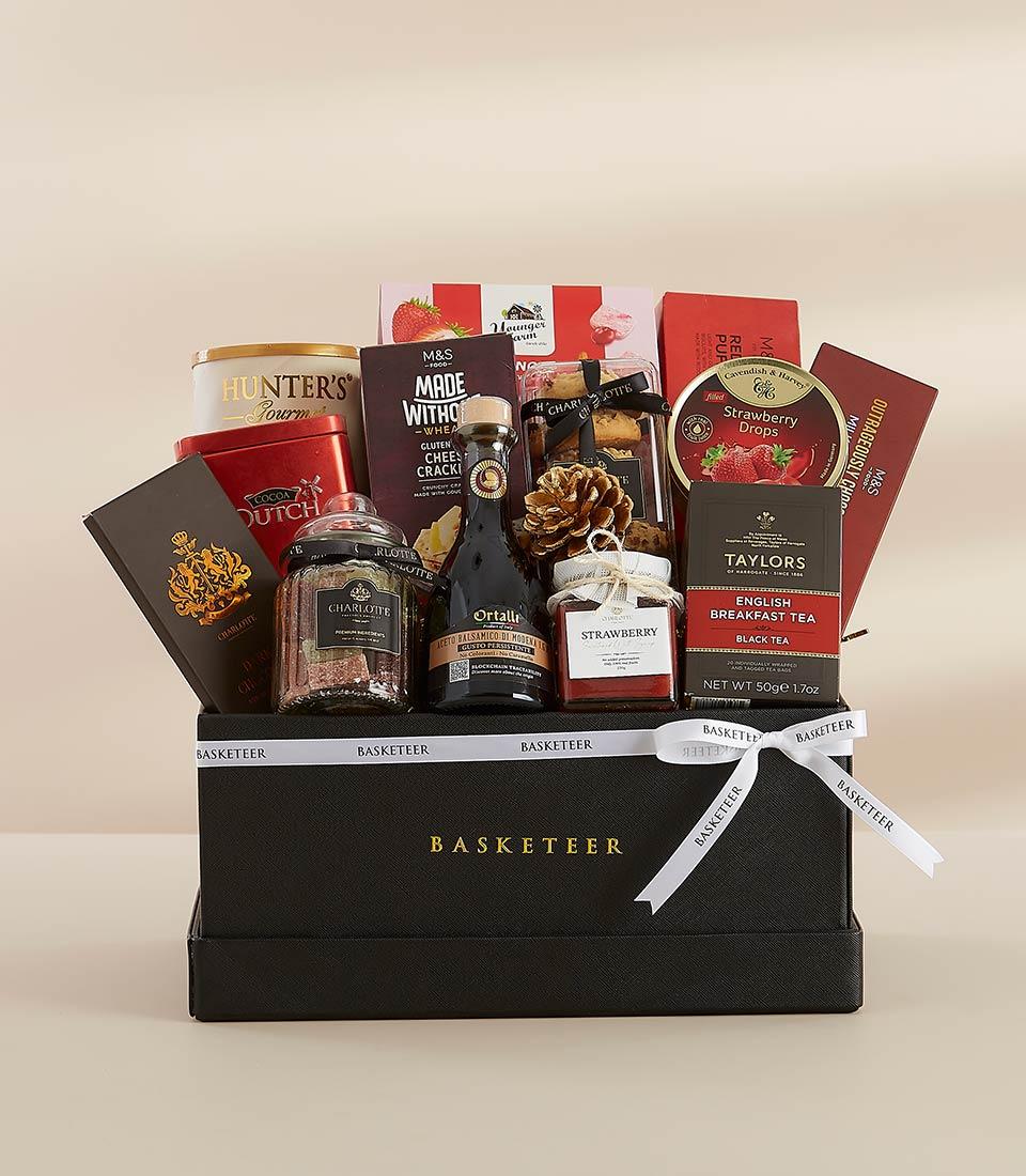 Black gift box filled with premium treats including gluten-free crackers, strawberry jam, English breakfast tea, Dutch cocoa, and wine bottle, tied with a white ribbon.