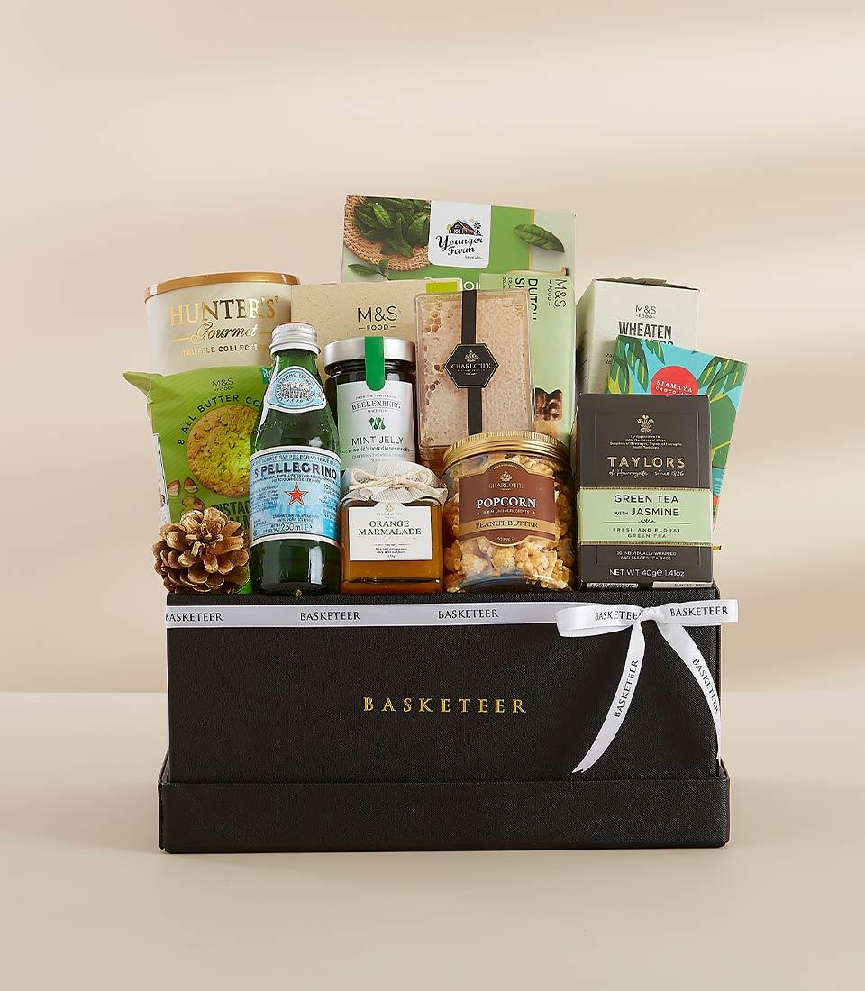 Elegant black gift box with green tea, San Pellegrino, orange marmalade, mint jelly, peanut butter popcorn, and savory snacks, tied with a white ribbon.