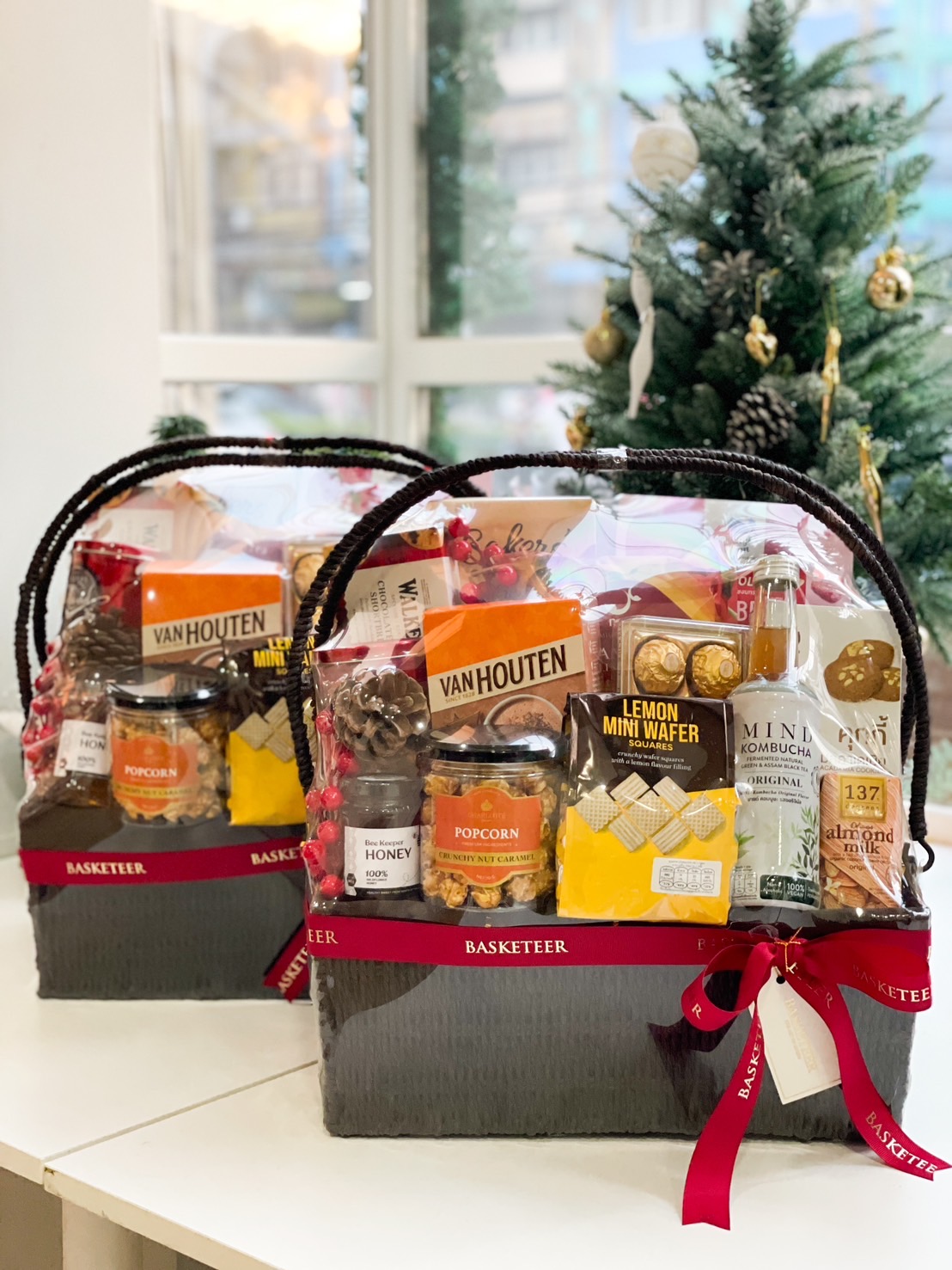 Elegant black gift basket filled with Van Houten chocolate, caramel popcorn, honey, lemon wafers, kombucha, almond milk, and other gourmet treats, adorned with Basketeer’s red ribbon.