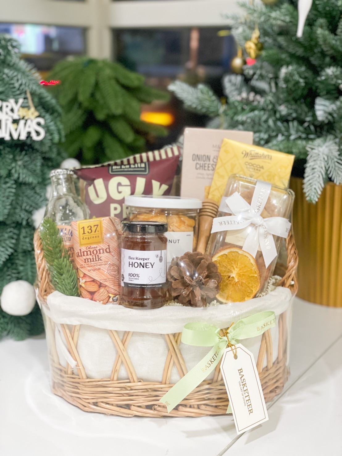 A wicker basket filled with honey, almond milk, onion & cheese crackers, cocoa blocks, and snacks, elegantly decorated with a green ribbon and pinecone.