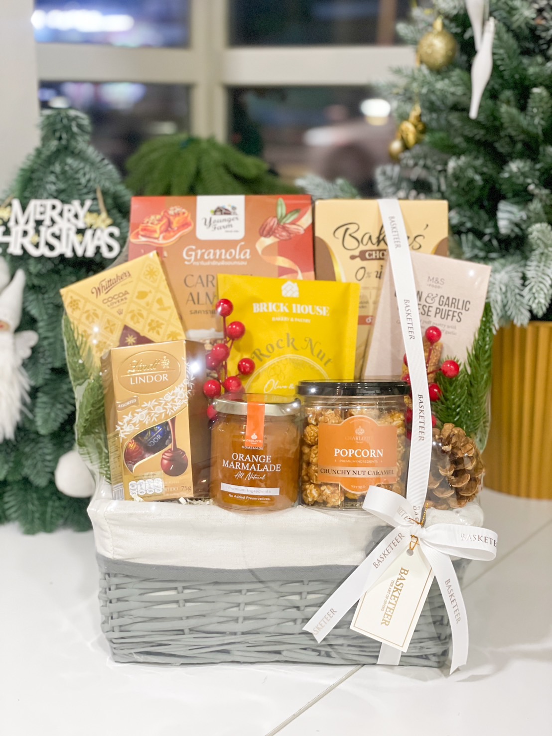 A grey wicker basket filled with chocolates, granola, marmalade, popcorn, and snacks, adorned with red berries and a white ribbon.