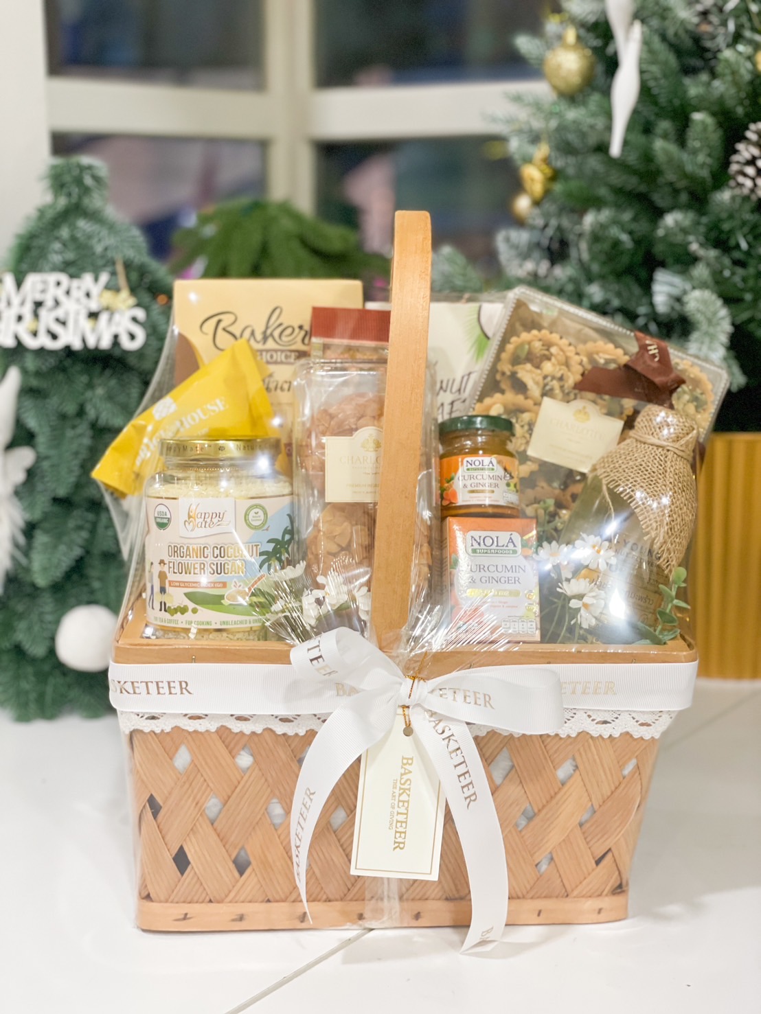 A beautifully woven wicker basket filled with organic tea, coconut sugar, granola, nuts, cookies, and honey, adorned with white lace and a Basketeer ribbon.