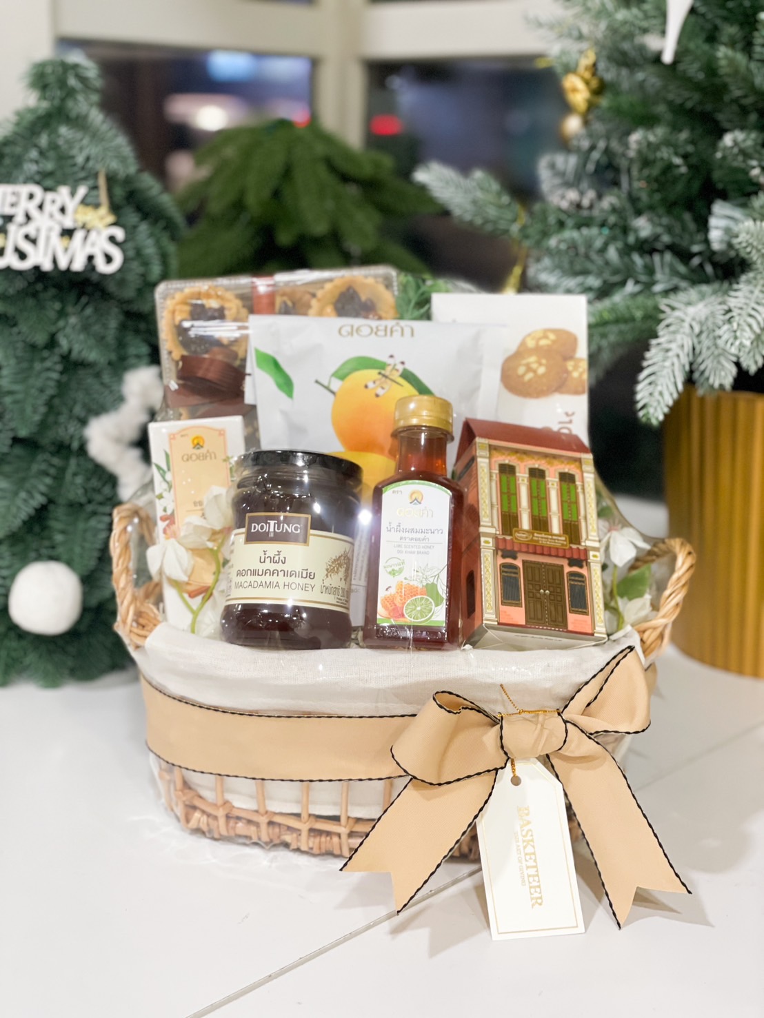 A beige wicker basket containing macadamia honey, lime honey syrup, cookies, and snacks, adorned with a beige ribbon and Basketeer tag.