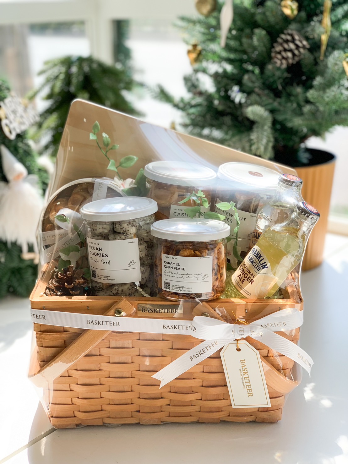 Artisanal gift basket featuring jars of vegan cookies, caramel cornflakes, cashew nuts, and a bottle of ginger ale, wrapped in clear covering and adorned with Basketeer ribbons.