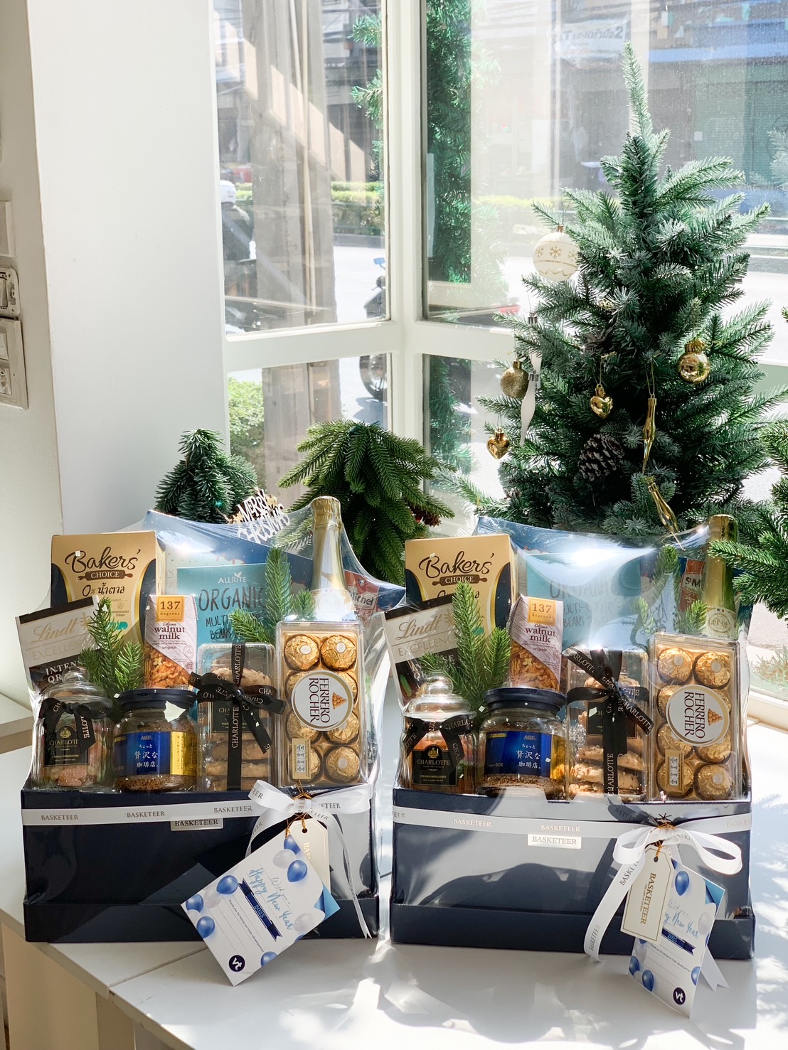 Two luxury holiday gift sets wrapped in clear packaging, containing chocolates, gourmet snacks, and festive decorations, placed on a white surface with a Christmas tree in the background.