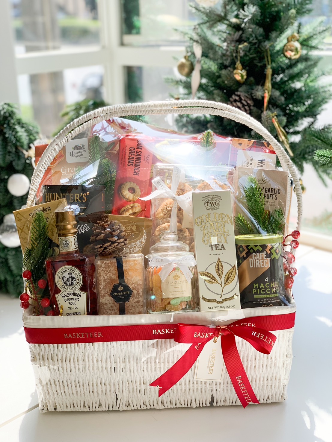 White wicker gift basket filled with premium snacks, teas, and condiments, decorated with festive accents, displayed in front of a Christmas tree.