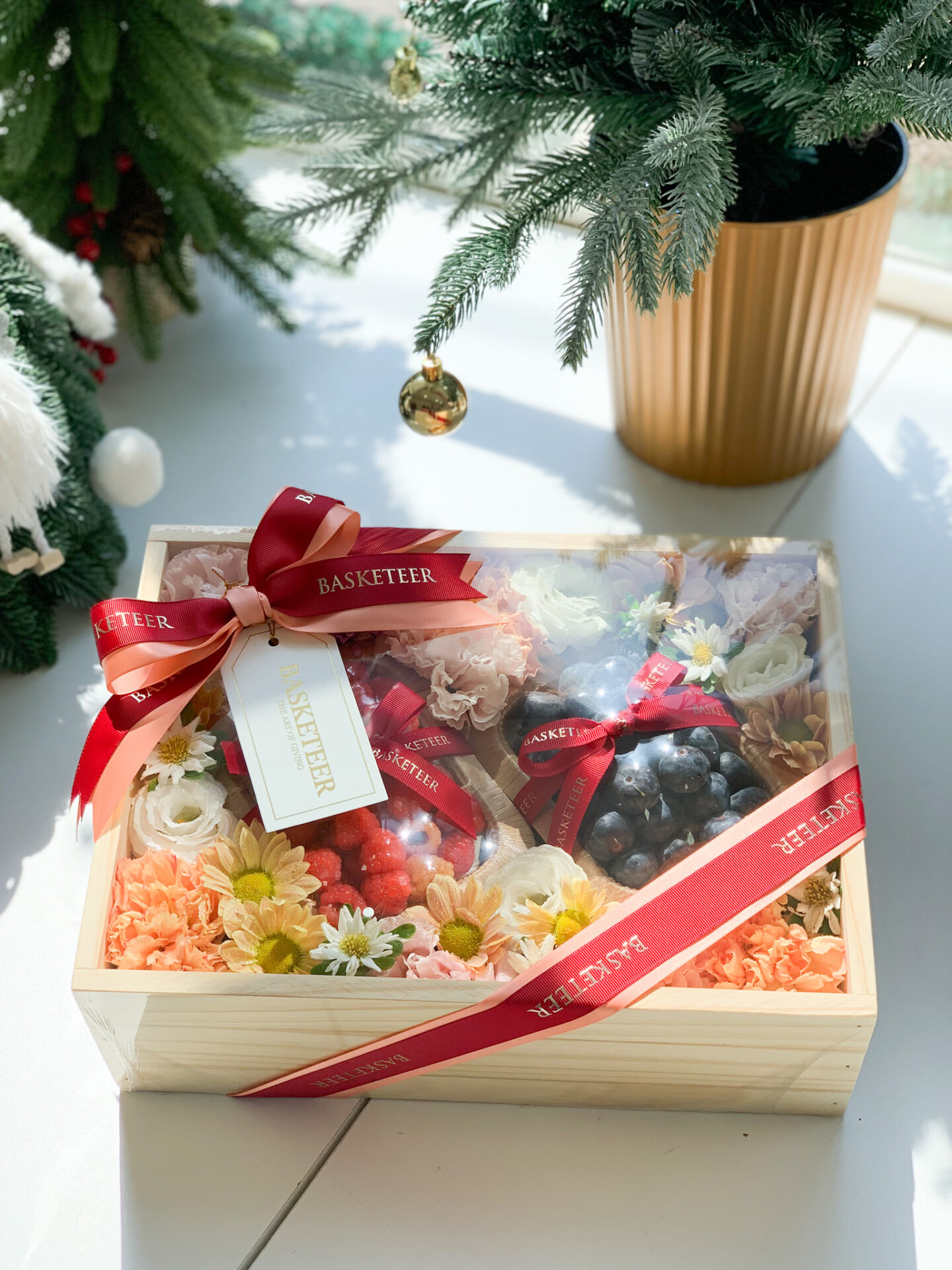 Wooden fruit box adorned with fresh flowers, berries, and wrapped with a red ribbon, displayed next to a festive Christmas tree.