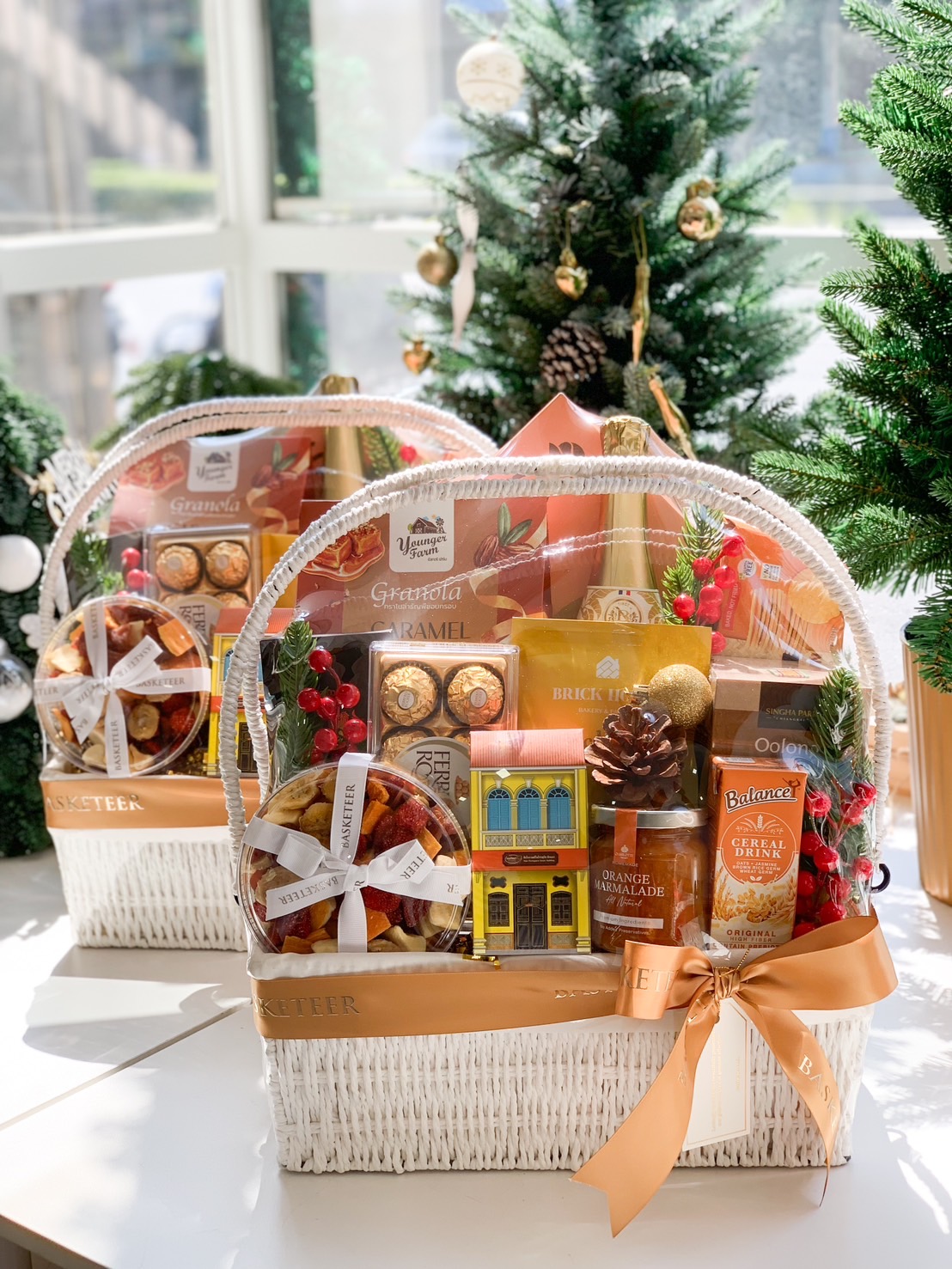 Elegant white wicker gift basket filled with Ferrero Rocher chocolates, marmalade, granola, festive decor, and tied with a golden ribbon, displayed in front of a Christmas tree.