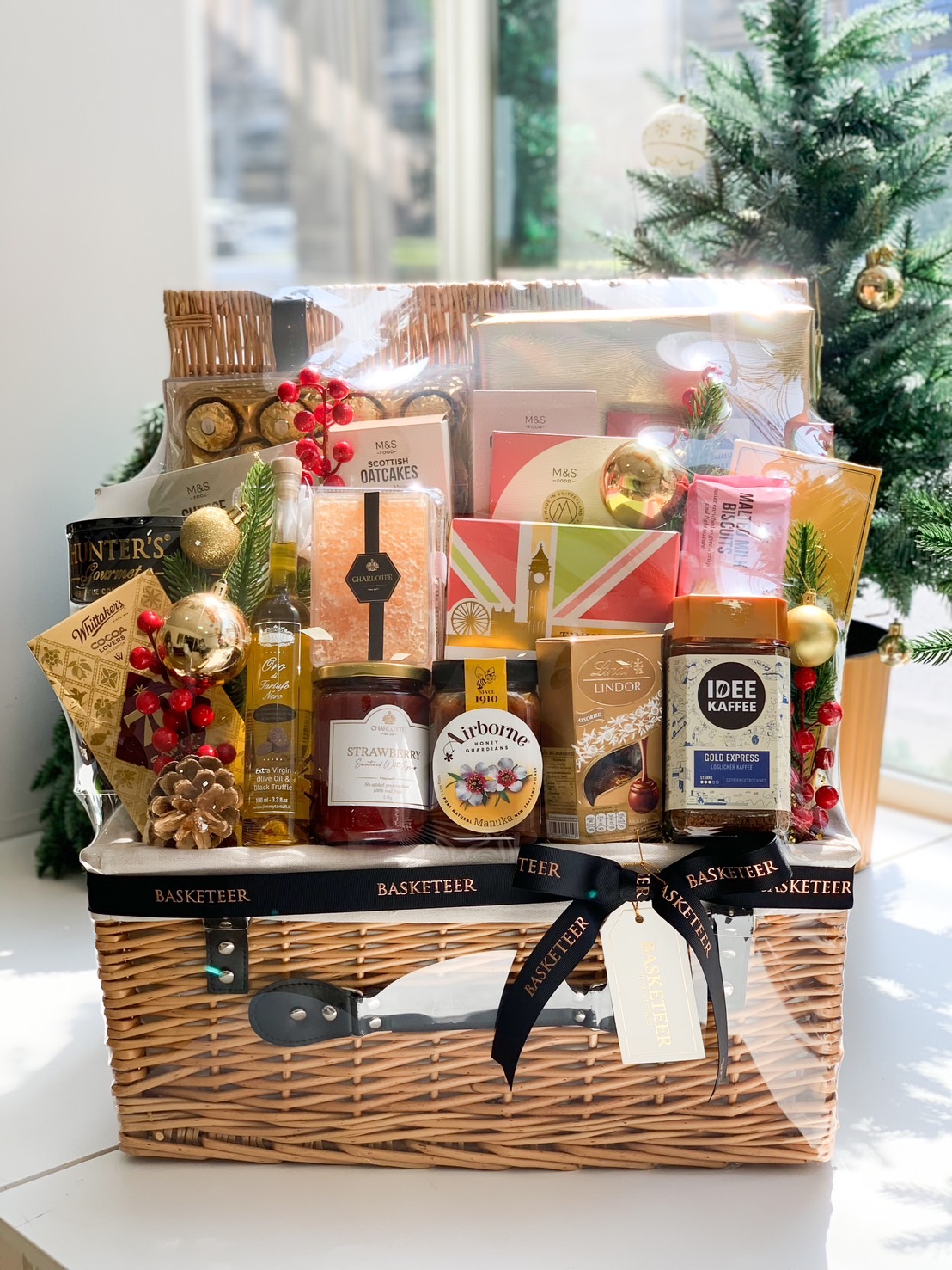Deluxe gift basket featuring premium chocolates, coffee, jam, honey, biscuits, and festive decorations, tied with a black ribbon and gold accents.