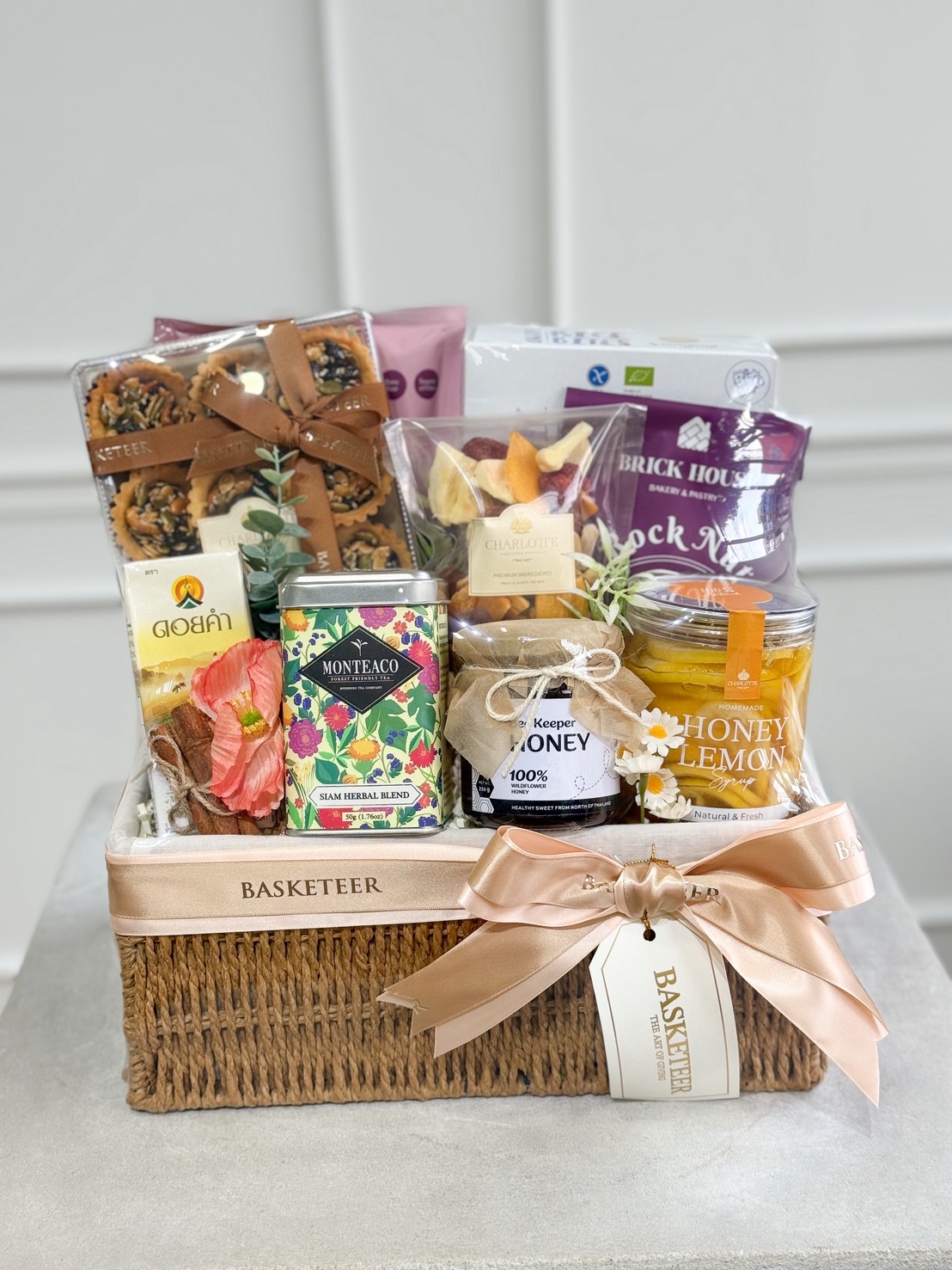 A refined wicker gift hamper with a beige ribbon, filled with premium healthy Thai snacks, tea, honey, and dried fruit.