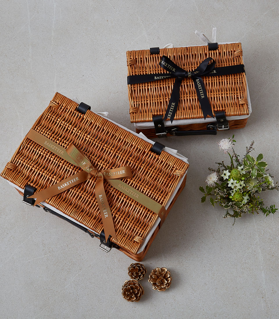 Two wicker gift baskets with ribbons, one larger and one smaller, are placed on a light grey surface. A small bouquet of wildflowers and three pine cones are arranged nearby, creating a scene that captures the essence of Peaceful Moments Gift.