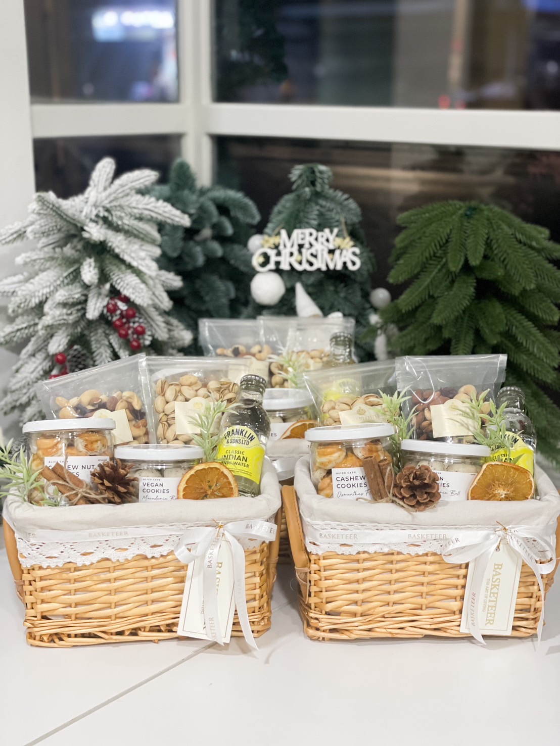 The Relaxation Station Gift Basket is a delightful wicker collection of assorted snacks, featuring cashew nut cookies, mixed nuts, and a refreshing bottle of lemonade. It sits elegantly on a marble countertop, wrapped with a ribbon and greenery against a tiled wall.
