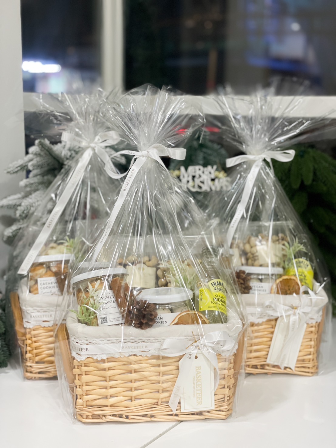The Relaxation Station Gift Basket is a delightful wicker collection of assorted snacks, featuring cashew nut cookies, mixed nuts, and a refreshing bottle of lemonade. It sits elegantly on a marble countertop, wrapped with a ribbon and greenery against a tiled wall.