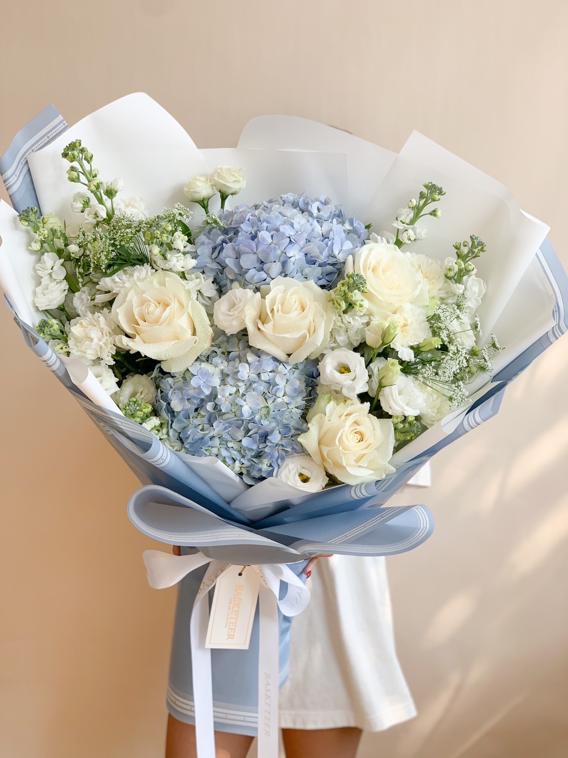 A stunning bouquet of blue hydrangeas and white roses, elegantly wrapped in soft blue paper with a Basketeer ribbon and tag, held by a person in a white outfit.
