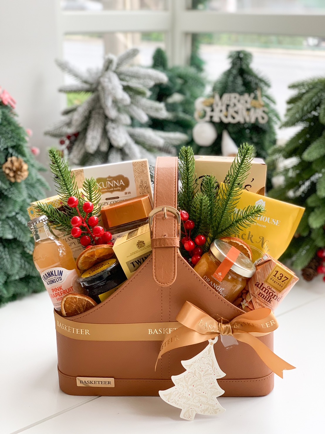 A gift basket filled with various gourmet items, such as jars, packets, and bottles, neatly arranged in a leather carrier with a ribbon. It’s decorated with sprigs of greenery and dried citrus. A flowering plant in a vase is in the background.