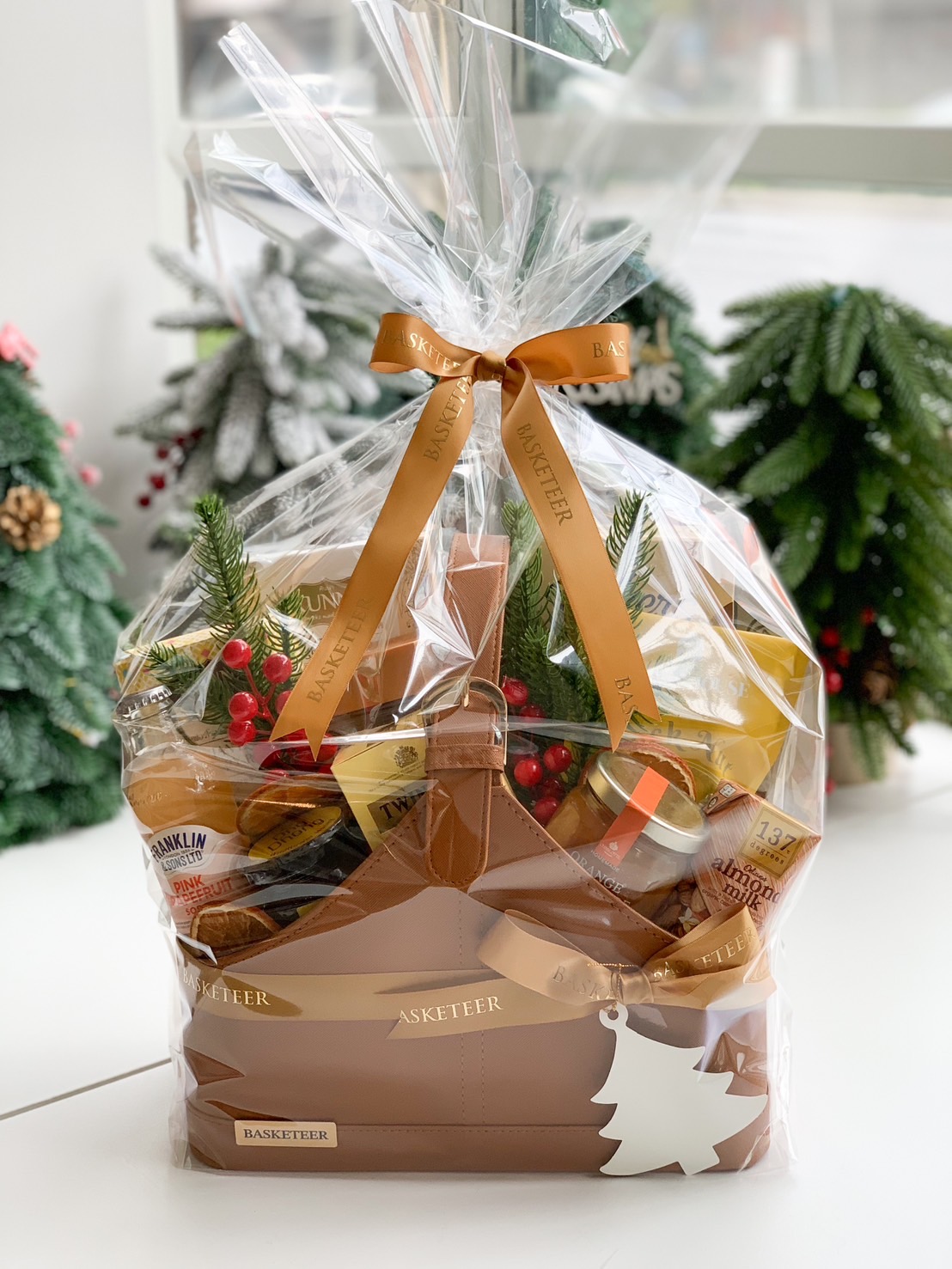 A gift basket filled with various gourmet items, such as jars, packets, and bottles, neatly arranged in a leather carrier with a ribbon. It’s decorated with sprigs of greenery and dried citrus. A flowering plant in a vase is in the background.