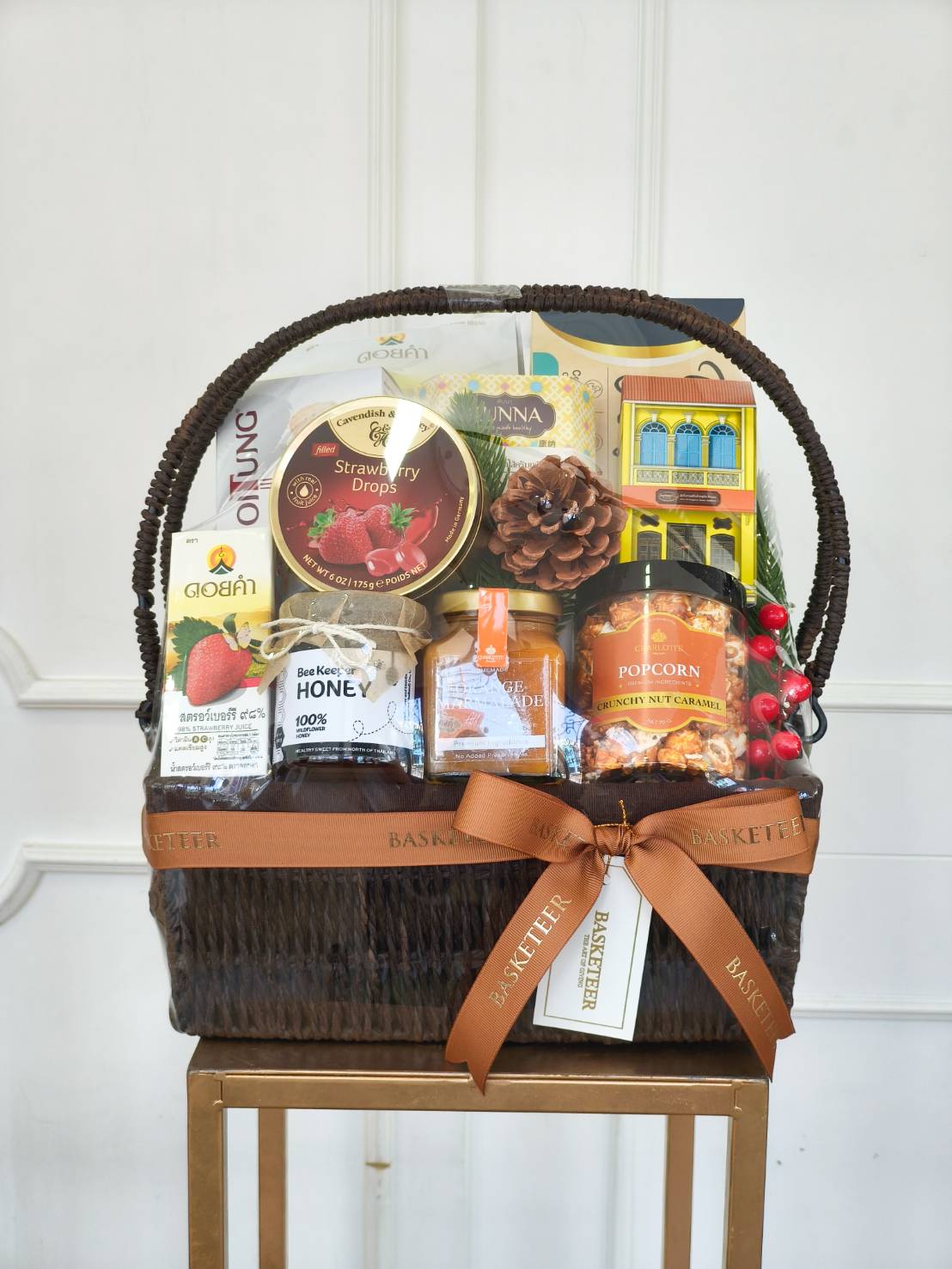 Luxury gourmet gift basket with premium snacks, honey, tea, strawberry drops, and caramel popcorn, elegantly wrapped with brown ribbon.